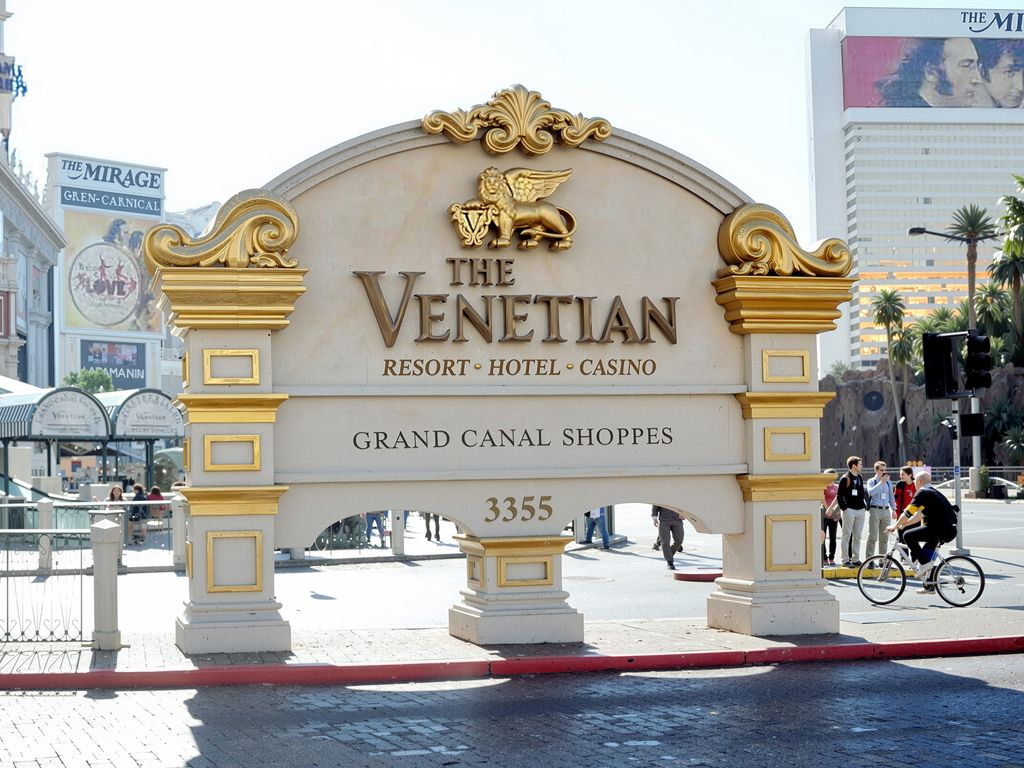 A sign for The Venetian Resort Hotel Casino in Las Vegas, featuring ornate gold accents and the address '3355'. A cyclist rides past the sign on the street.